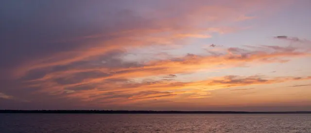 Layered orange and violet sunset clouds stretch over calm bay water and a distant tree-lined shore