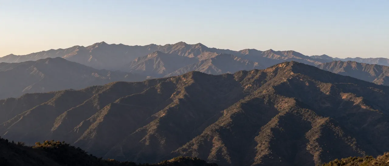 Layered mountain peaks stretch across the view as warm sunrise light falls on ridgelines under a pale sky