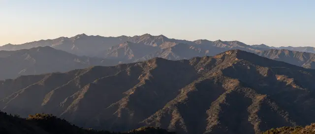 Layered mountain peaks stretch across the view as warm sunrise light falls on ridgelines under a pale sky