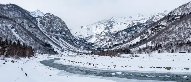 Overcast panoramic view of a snowy mountain valley with a partially frozen winding river and surrounding snow-capped peaks