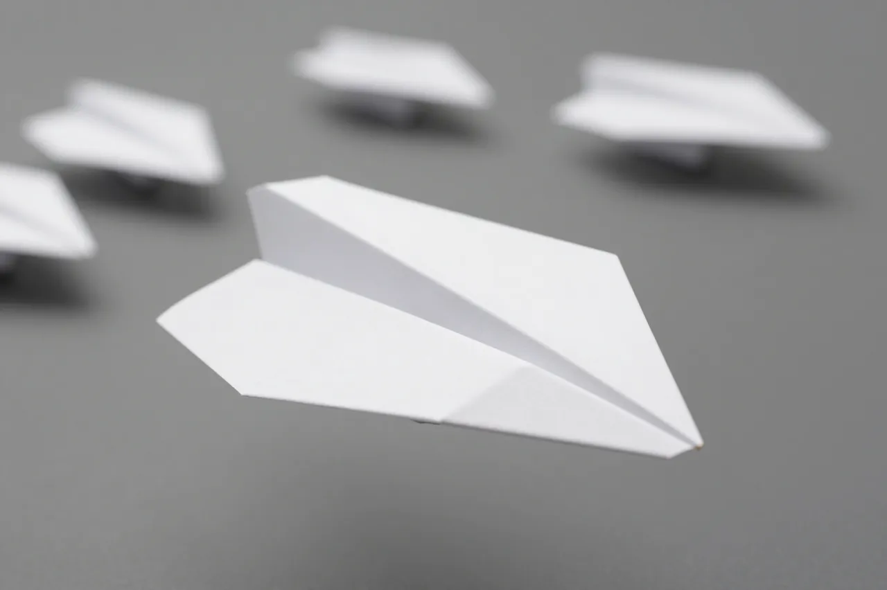 A sharp foreground white paper airplane leads a blurred line of other paper airplanes above a plain gray surface