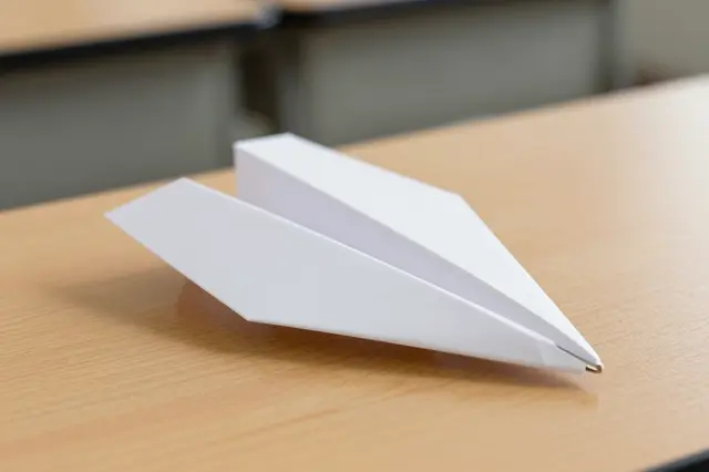 A white folded paper airplane rests on a light wooden classroom desk with blurred desks in the background.