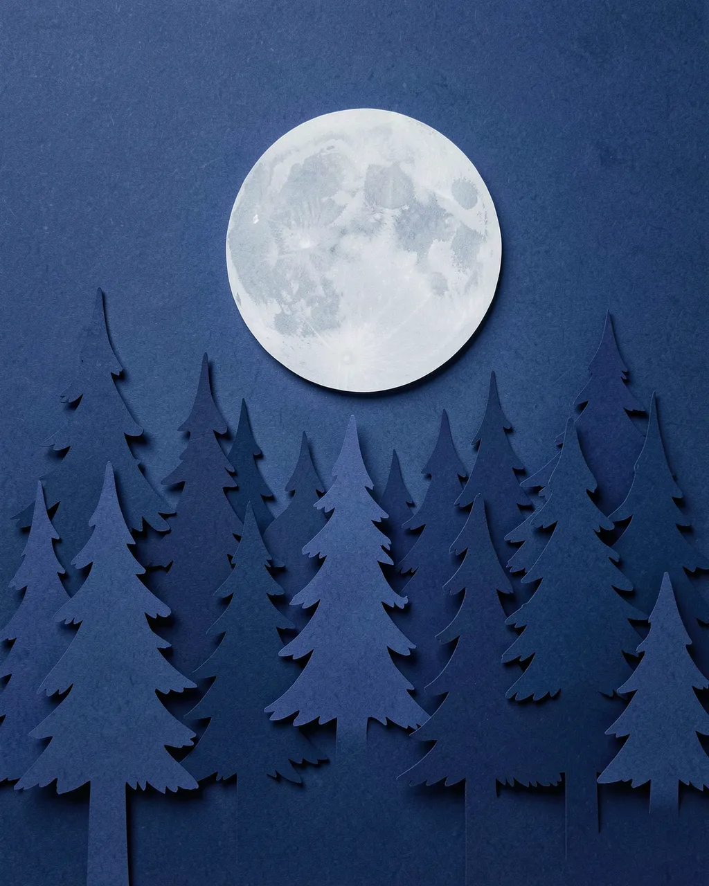 Layered paper cut art of a pine forest beneath a bright full moon on a midnight blue background