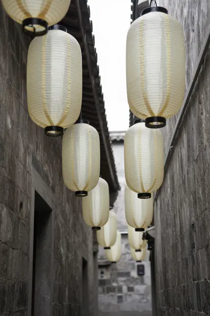 Rows of plain white paper lanterns hang along a narrow stone old town alley under overcast sky