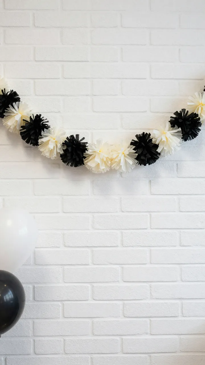 Black and cream tissue pom pom garland hangs above balloons against a white painted brick wall with empty copy space.