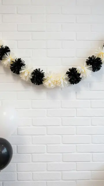 Black and cream tissue pom pom garland hangs above balloons against a white painted brick wall with empty copy space.
