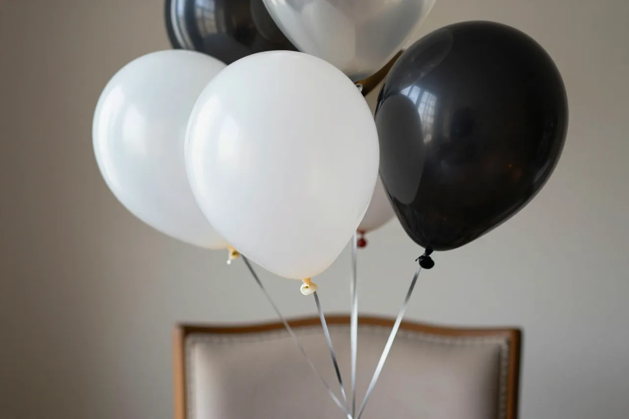 Black, white and clear helium balloons are tied to the back of an upholstered wooden chair against a plain wall