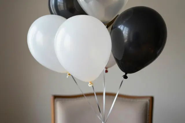 Black, white and clear helium balloons are tied to the back of an upholstered wooden chair against a plain wall