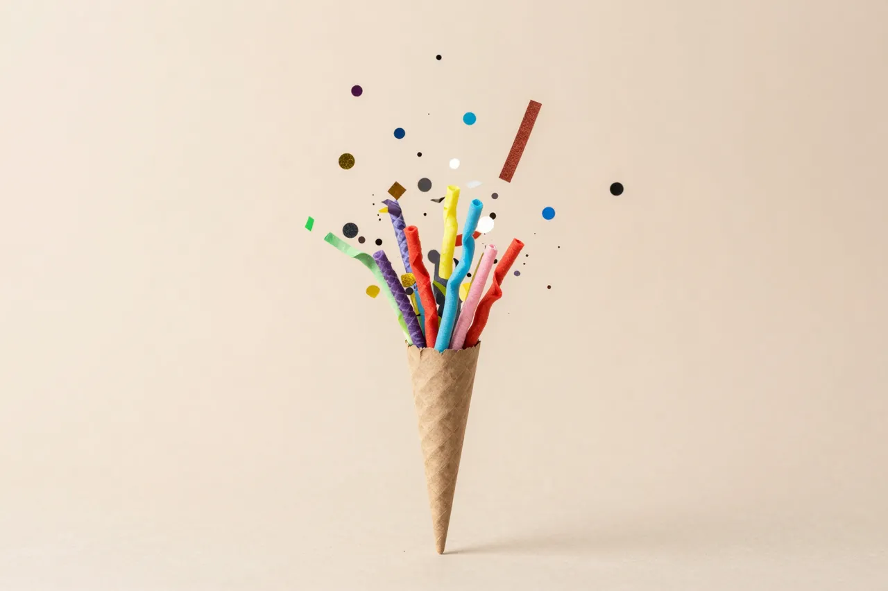 Colorful confetti and streamers burst upward from a kraft paper cone against a soft beige background