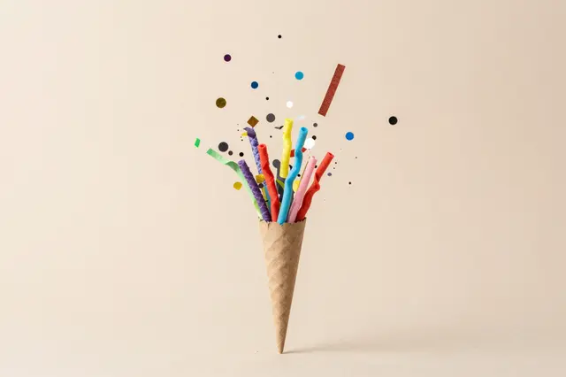 Colorful confetti and streamers burst upward from a kraft paper cone against a soft beige background