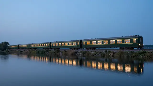 Lit passenger train runs along a still lakeshore with glowing window reflections at blue hour