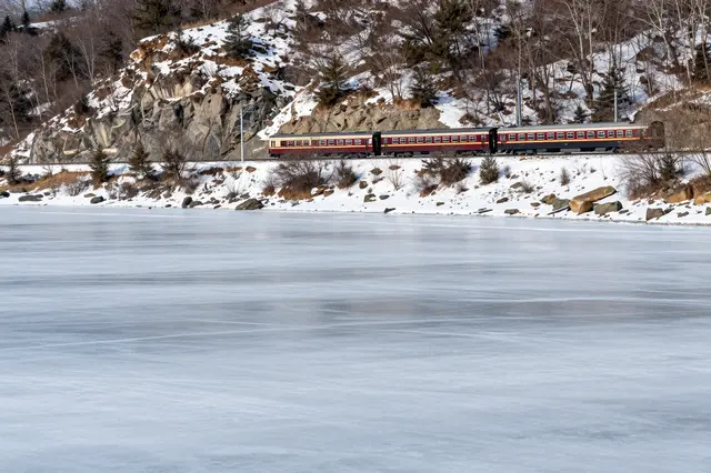 A passenger train travels along a snow-dusted railway beside a frozen lake under rocky winter hills