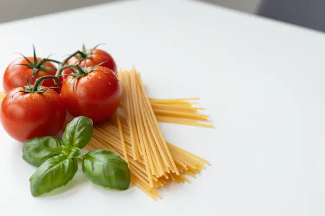 Fresh vine tomatoes, basil and dry spaghetti rest on the left of a plain white table with empty copy space to the right