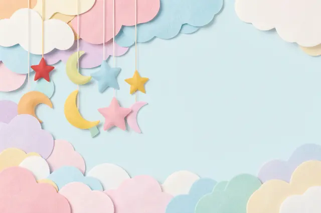 Felt star and moon baby mobile hangs over layered pastel clouds with blank copy space on a light blue background.