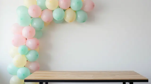 A pastel balloon garland arcs above an empty wooden table against a plain white wall with right-side copy space.