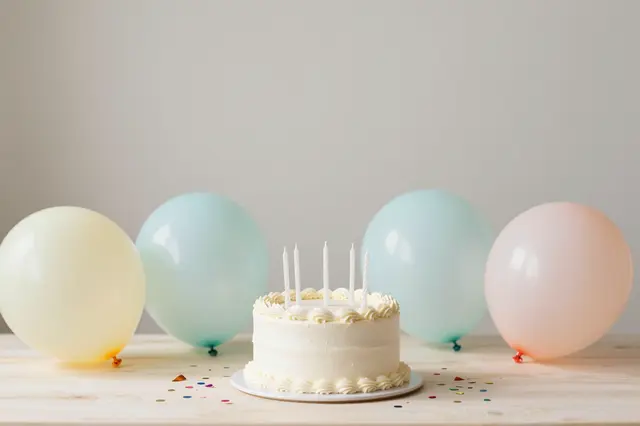 White frosted birthday cake sits on a light wooden table surrounded by pastel balloons and scattered confetti with empty