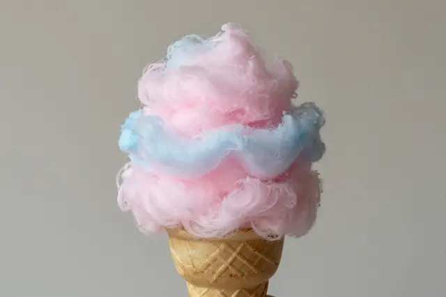 Pink and blue swirled cotton candy rests in a waffle cone against a plain light gray background