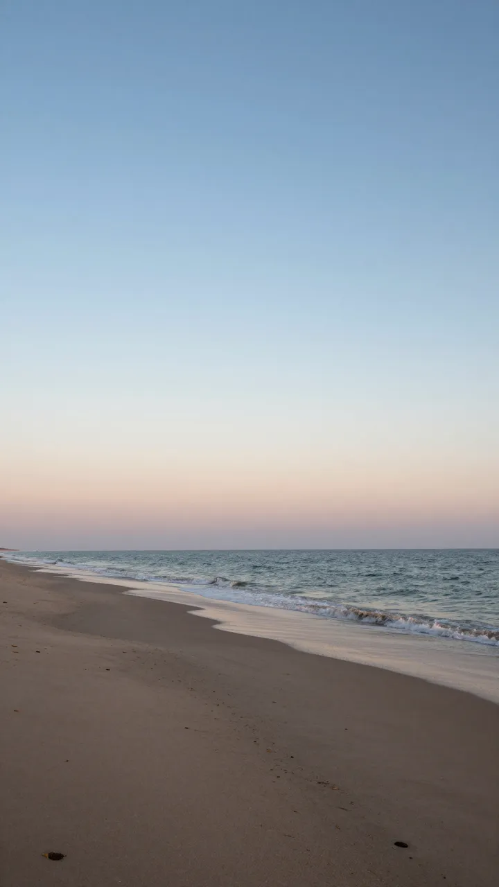 A quiet sandy beach stretches beside gentle ocean waves under a soft pastel dawn sky.