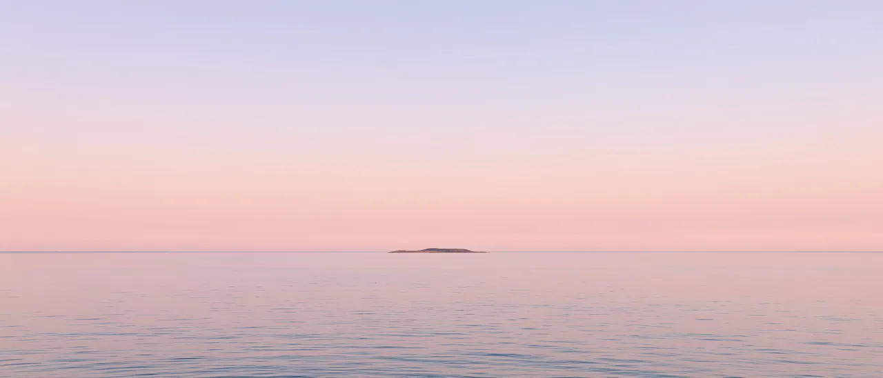 Wide view of a calm flat sea with a tiny distant island under a soft pastel gradient dawn sky