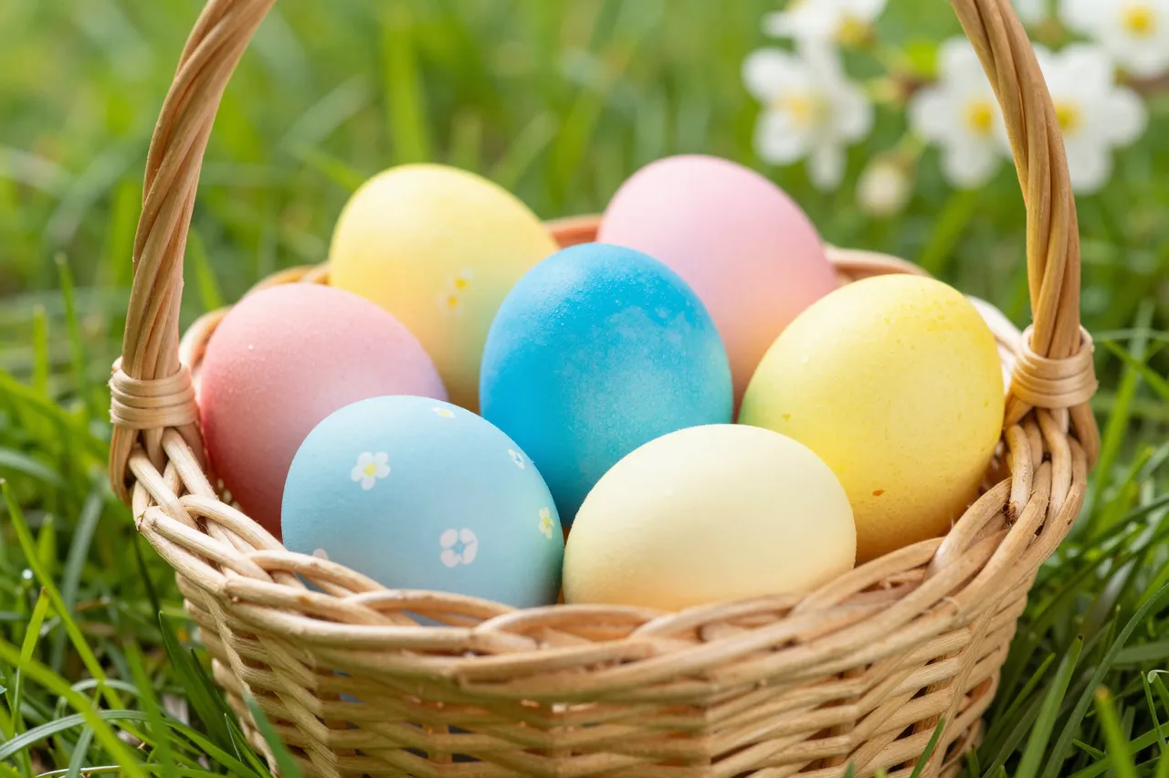 A wicker basket filled with decorated pastel Easter eggs rests on lush green spring grass with blurred white flowers in the