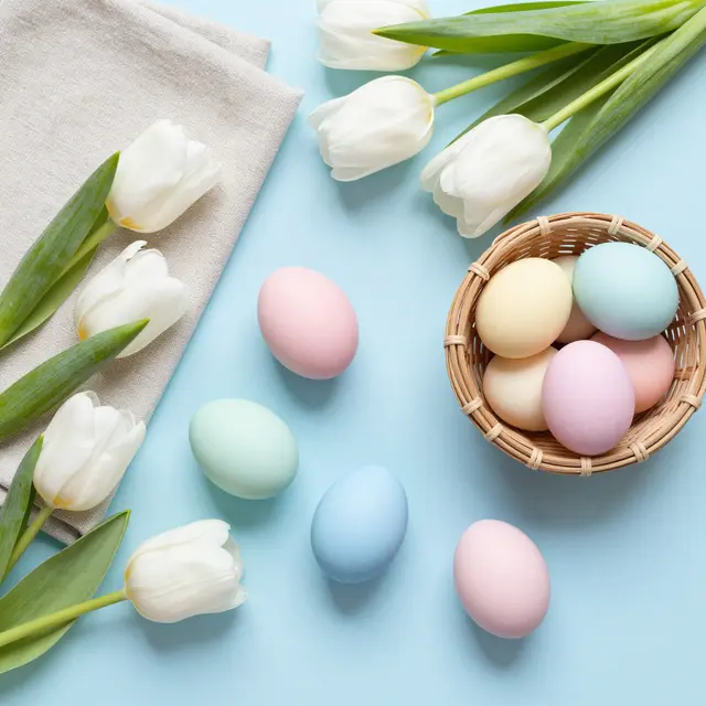 Top-down flat lay of pastel Easter eggs, white tulips, linen napkin and wicker basket on pale blue background