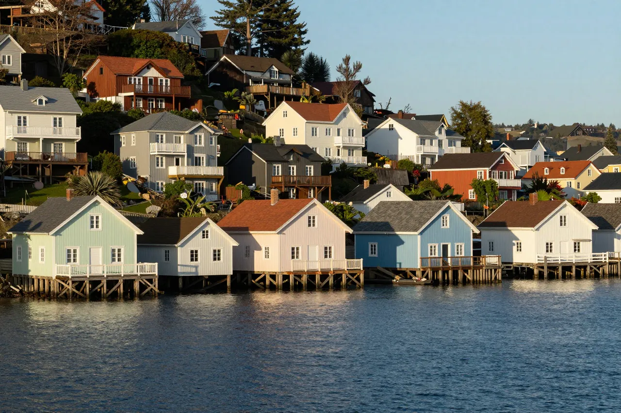 Stilted pastel waterfront houses line calm blue water under warm morning sunlight on a sloping harbor hillside