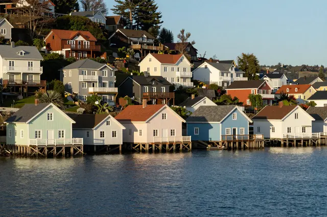 Stilted pastel waterfront houses line calm blue water under warm morning sunlight on a sloping harbor hillside