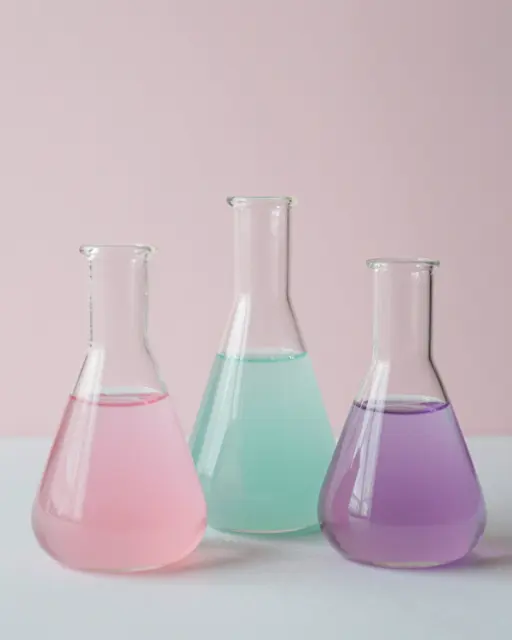 Three glass erlenmeyer flasks holding pink, mint, and lilac liquid sit on a pale surface against a soft pink wall
