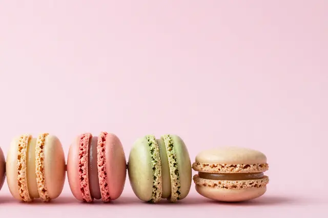 Row of assorted pastel macarons arranged along the bottom edge of a solid soft pink background with empty copy space above