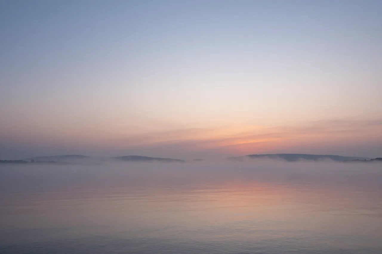 Soft pastel sunrise hues reflect on calm misty bay water with distant hazy shorelines