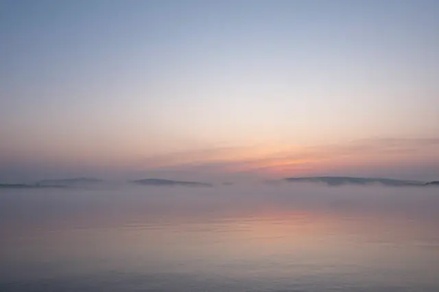 Soft pastel sunrise hues reflect on calm misty bay water with distant hazy shorelines