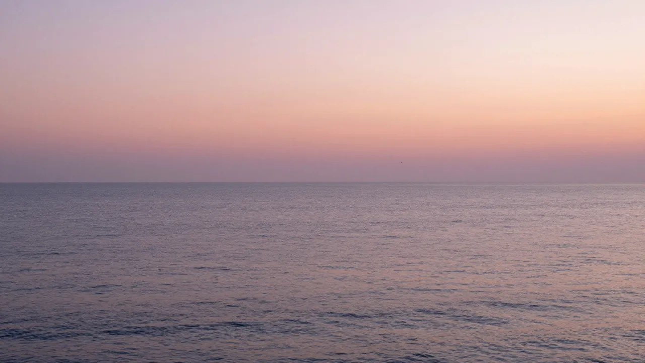 Calm still ocean stretches to the horizon beneath a soft pink and orange gradient sunrise sky