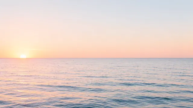 Low sun at the left horizon casts soft pastel light over calm, gentle ocean waves under an open sky