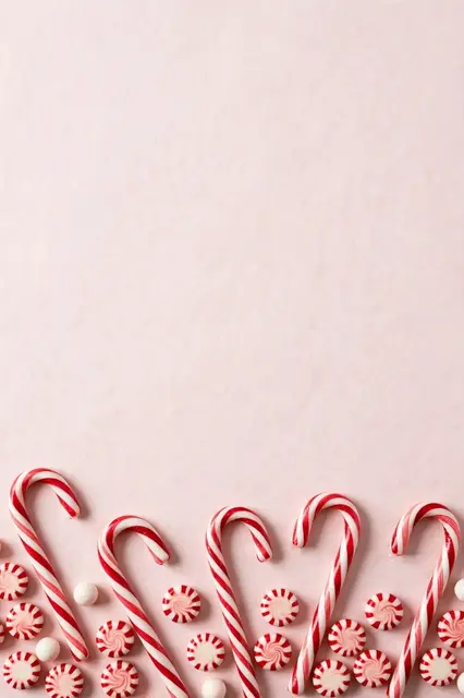 Overhead flat lay of red and white candy canes and peppermint candies arranged along the bottom edge of a soft pale pink