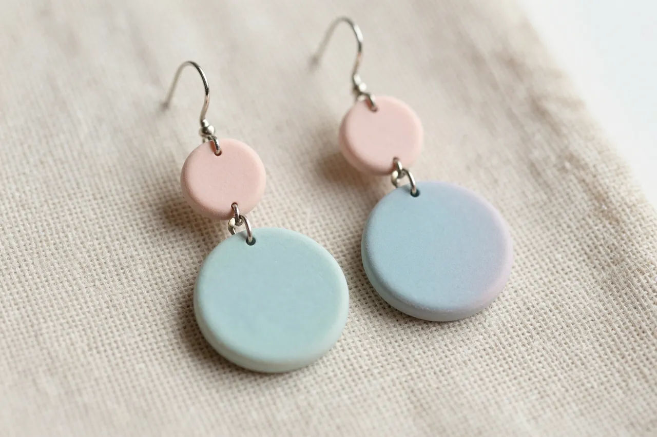 Dangle polymer clay earrings in soft pastel pink and blue rest on textured linen fabric.