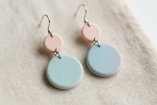 Dangle polymer clay earrings in soft pastel pink and blue rest on textured linen fabric.