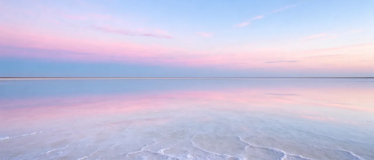 A still shallow salt lake perfectly mirrors a soft pink and blue gradient sky at dusk, with a thin distant horizon line