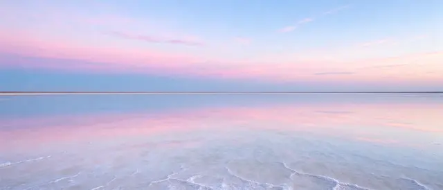 A still shallow salt lake perfectly mirrors a soft pink and blue gradient sky at dusk, with a thin distant horizon line