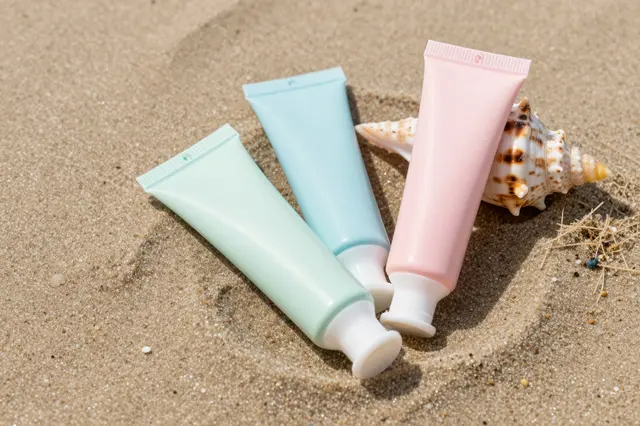 Three blank pastel squeeze tubes rest next to a spotted seashell on fine beach sand.