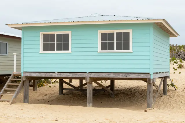 A light turquoise beach house elevated on wooden stilts sits on sandy beach under an overcast sky