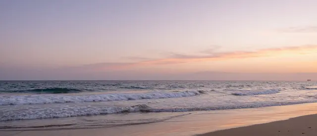 Gentle rolling waves wash onto a sandy shore under a soft gradient pastel sunset sky