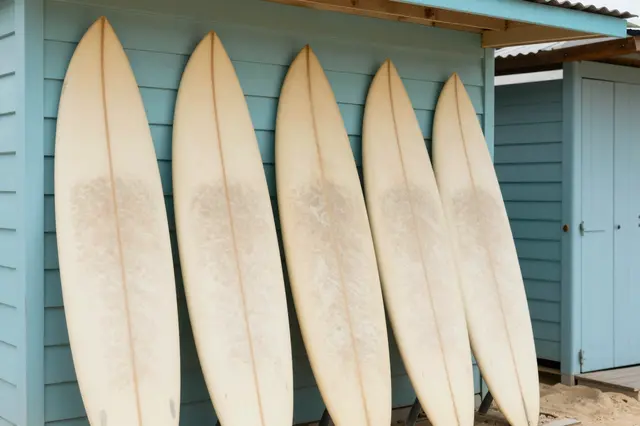 Five cream surfboards lean against a light turquoise wooden beach hut on sand