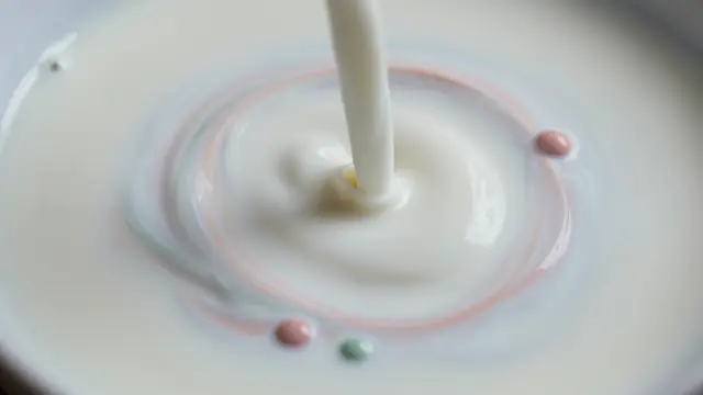 A stream of milk pours into a surface dotted with pastel pink and green food coloring drops forming soft swirling rings.