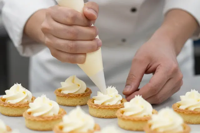 A pastry chef uses a piping bag to add cream to small tartlets on a work surface