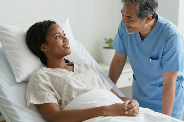 A smiling female patient rests in a hospital bed as a male nurse leans in to speak with her.