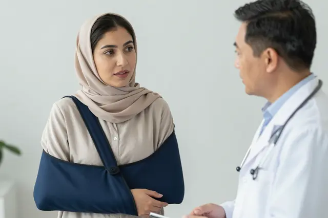 A woman with an arm sling speaks with an East Asian male doctor in a clinic consultation.