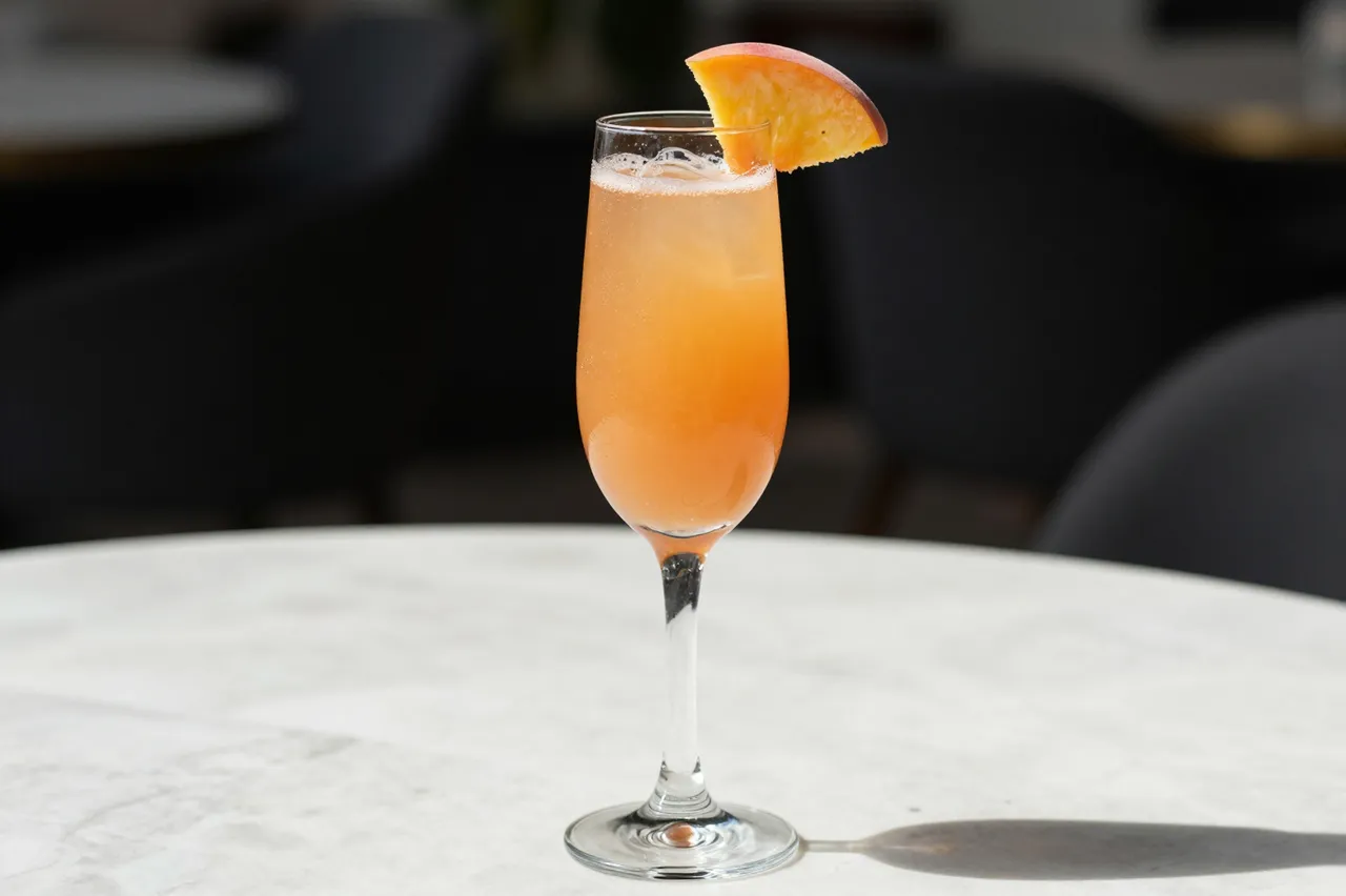 A tall stemmed flute of peach bellini with a fresh peach slice garnish sits on a sunlit marble table