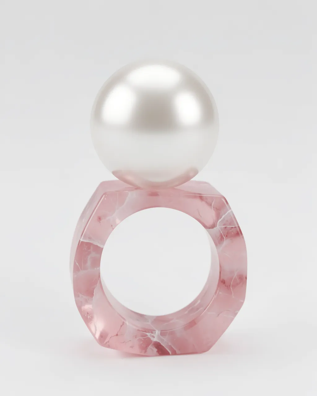 A smooth glossy pearl rests balanced on top of a faceted marbled rose quartz ring against a plain light gray background