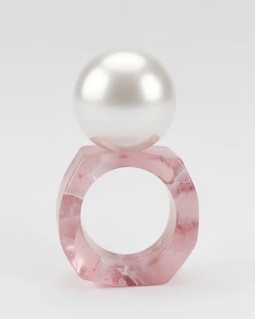 A smooth glossy pearl rests balanced on top of a faceted marbled rose quartz ring against a plain light gray background