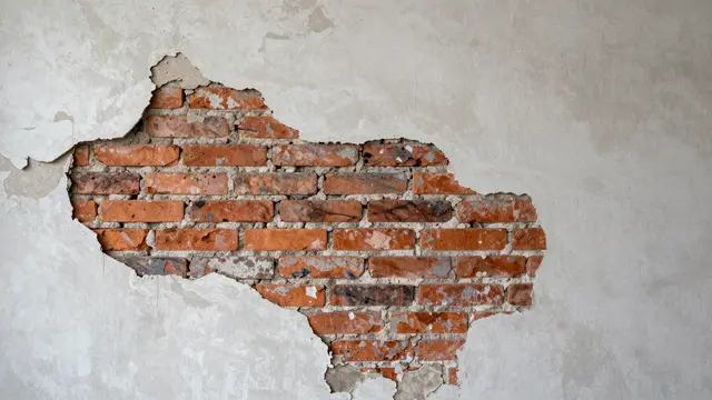 A wall section with peeling off-white plaster revealing weathered red brick underneath